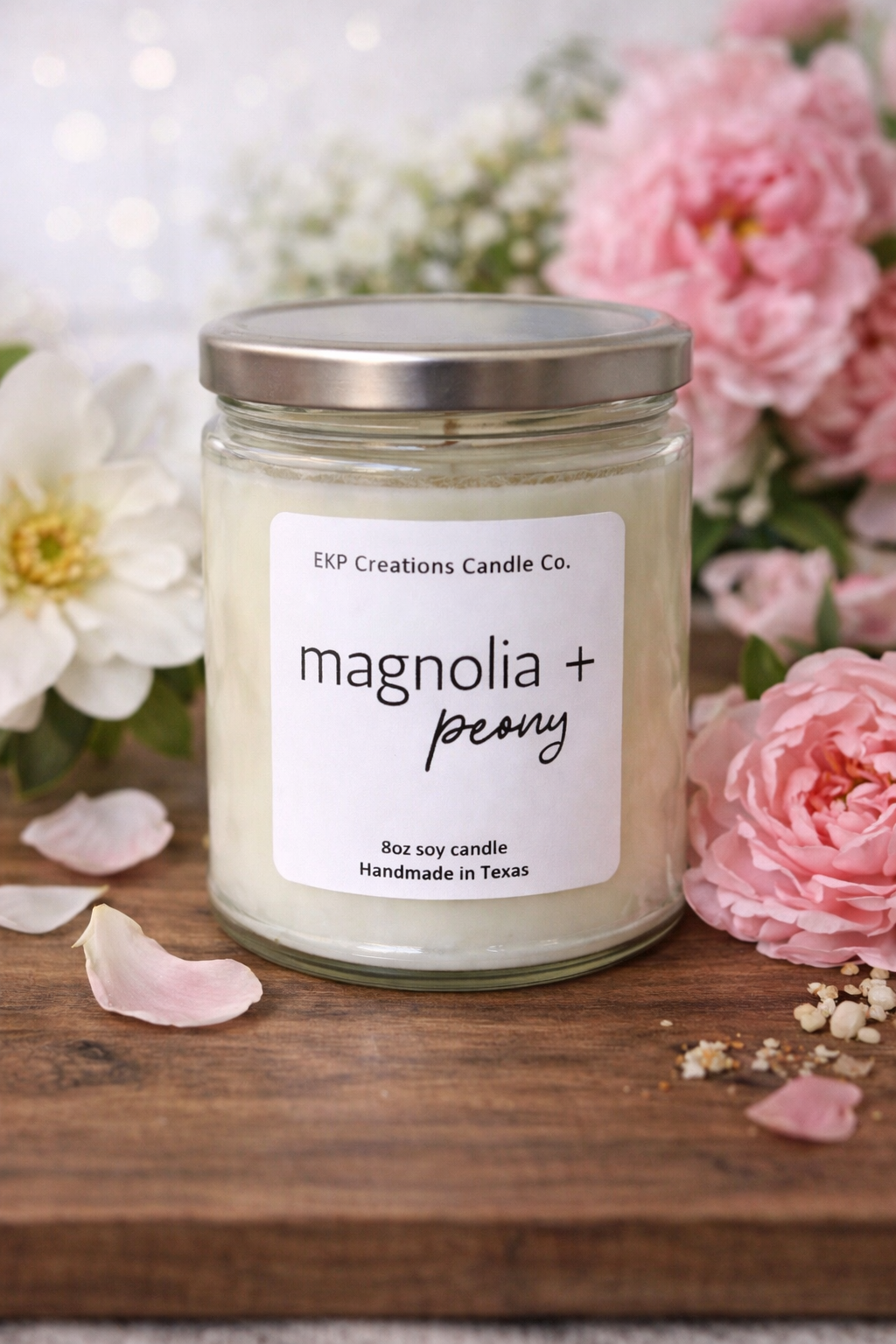 Candle jar labeled 'magnolia + peony' with flowers in the background. Scented soy candle handmade in Plano, Texas